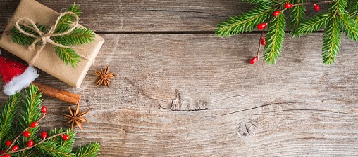 IMAGE DESCRIPTION: Picture shows a small gift wrapped in brown paper and tied with a jute string, pine branches with bright red berries nestled in them, a small red Santa’s cap and some spices. They are artistically laid out on a wood background