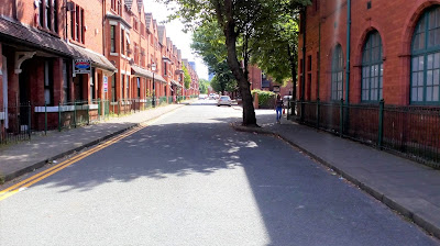 A street with buildings on both sides in old red brick. There is a large tree on the right which has a kerb around it to make a build out.