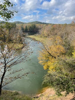 A photo looking out over a river. There's a few trees in the foreground that are losing their leaves for the fall. Then the river curving through fields and mountains. A sunny day.