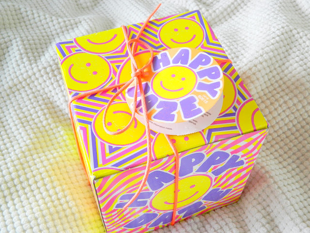 A photo showing the box of the Happy Daze gift set by Lush Cosmetics, featuring purple, pink and yellow design with smiley faces and stripes.