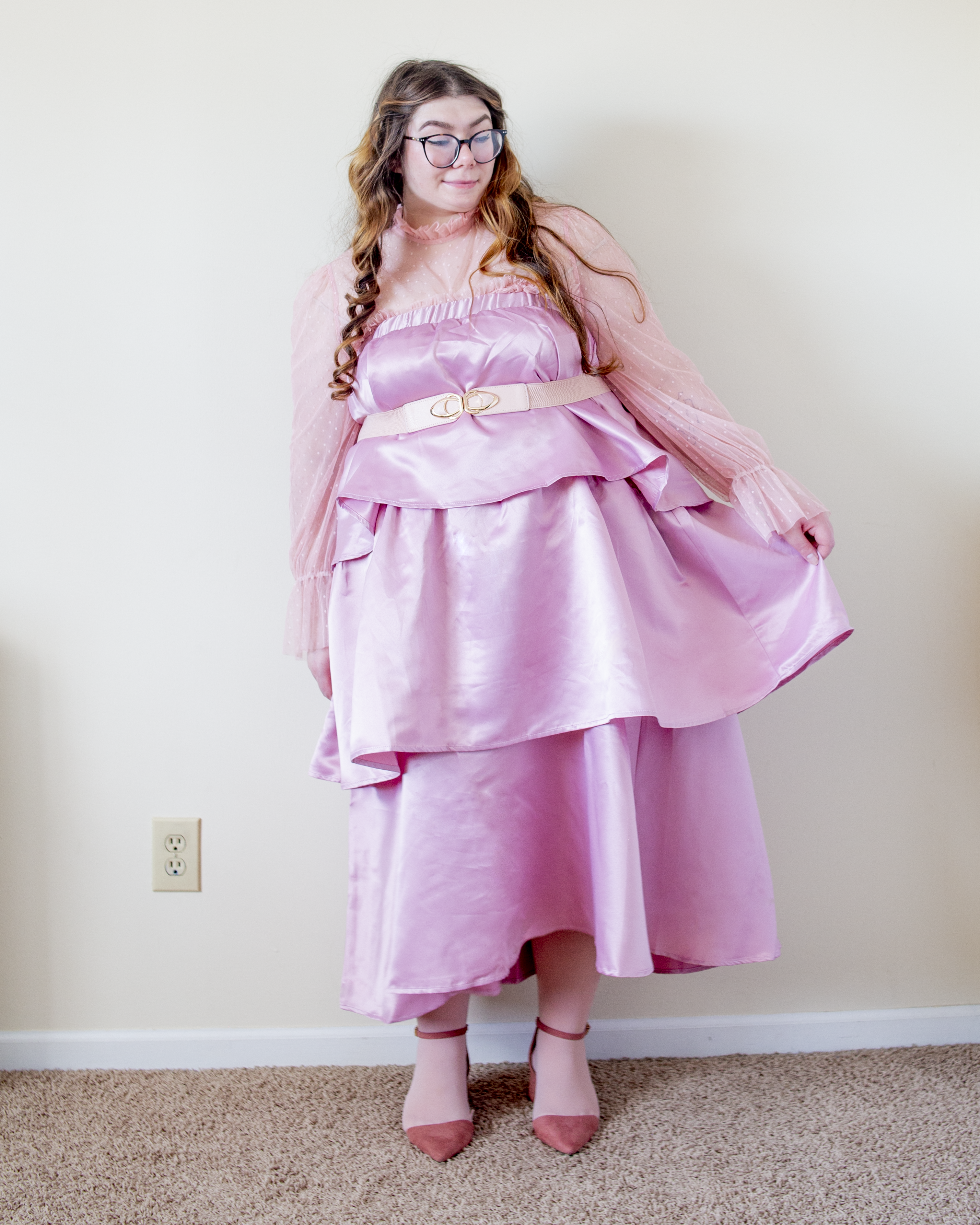 An outfit consisting of a pastel pink high neck long sleeve polka dot sheer blouse with a ruffle neckline under a three tiered pastel pink satin skirt, worn as dress, and pastel pink ankle strap heels.