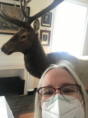 Woman with straight gray hair, glasses, and a white mask takes a selfie. Behind her is a taxidermied elk, a window with sun shining through, and several framed 18th or 19th century prints of different sizes.