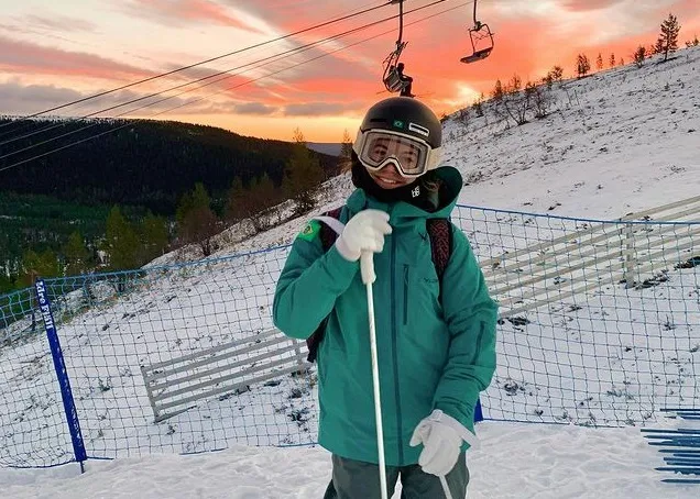 Sabrina posa para foto e sorri ao lado de pista de ski. Ela veste um casaco verde e capacete preto