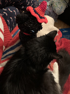 Viewed from behind and slightly above, Jinx is seated next to a much larger fluffy black and white cat. We see predominantly black in this photo. Both cats are facing the same direction; we don't see what they're looking at.