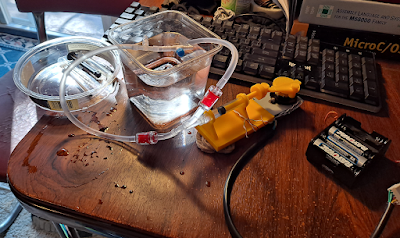 A little hydraulic syringe pump, battery pack, reservoir, "actuator" syringe, tubing, etc. sprawling across a card table with a keyboard in the background.