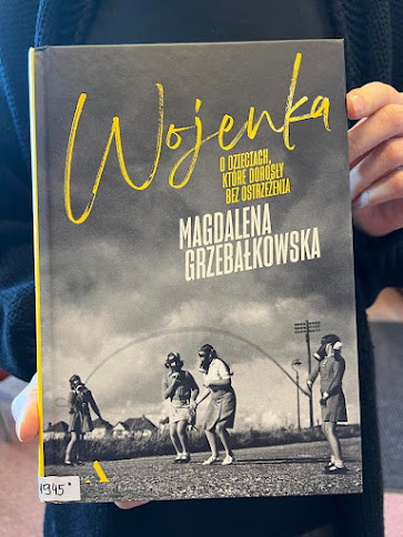 Czytelniczka w granatowym sweterku prezentuje książkę Magdaleny Grzebałkowskiej pod tytułem Wojenka: o dzieciach, które dorosły bez ostrzeżenia.