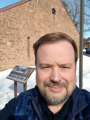 A man with brown hair, close-cut beard and mustache, and dark blue jacket takes a selfie. Behind him is snow-covered ground, a metal pole for a light fixture, and one wall of a brown stone building with a narrow window and a round window near the peak.