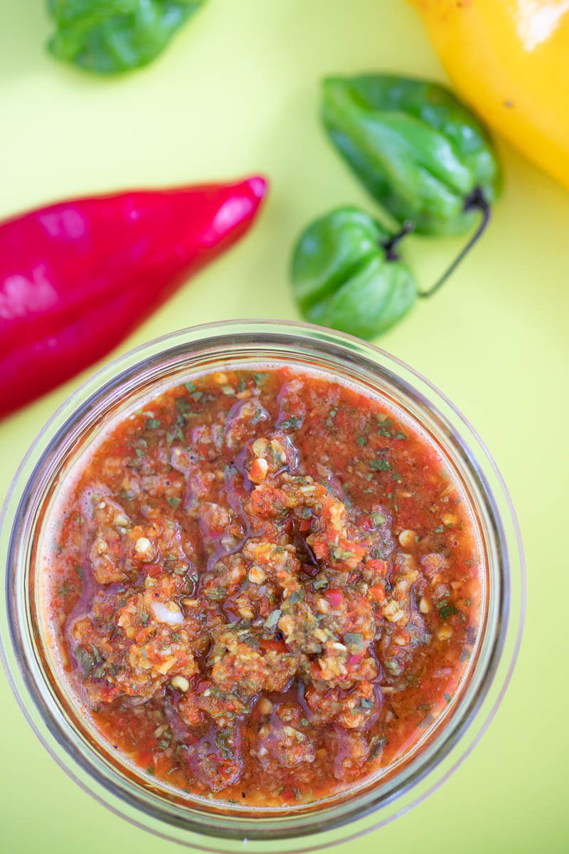 A close up of a jar of trinidad pepper sauce, with some peppers in the background.