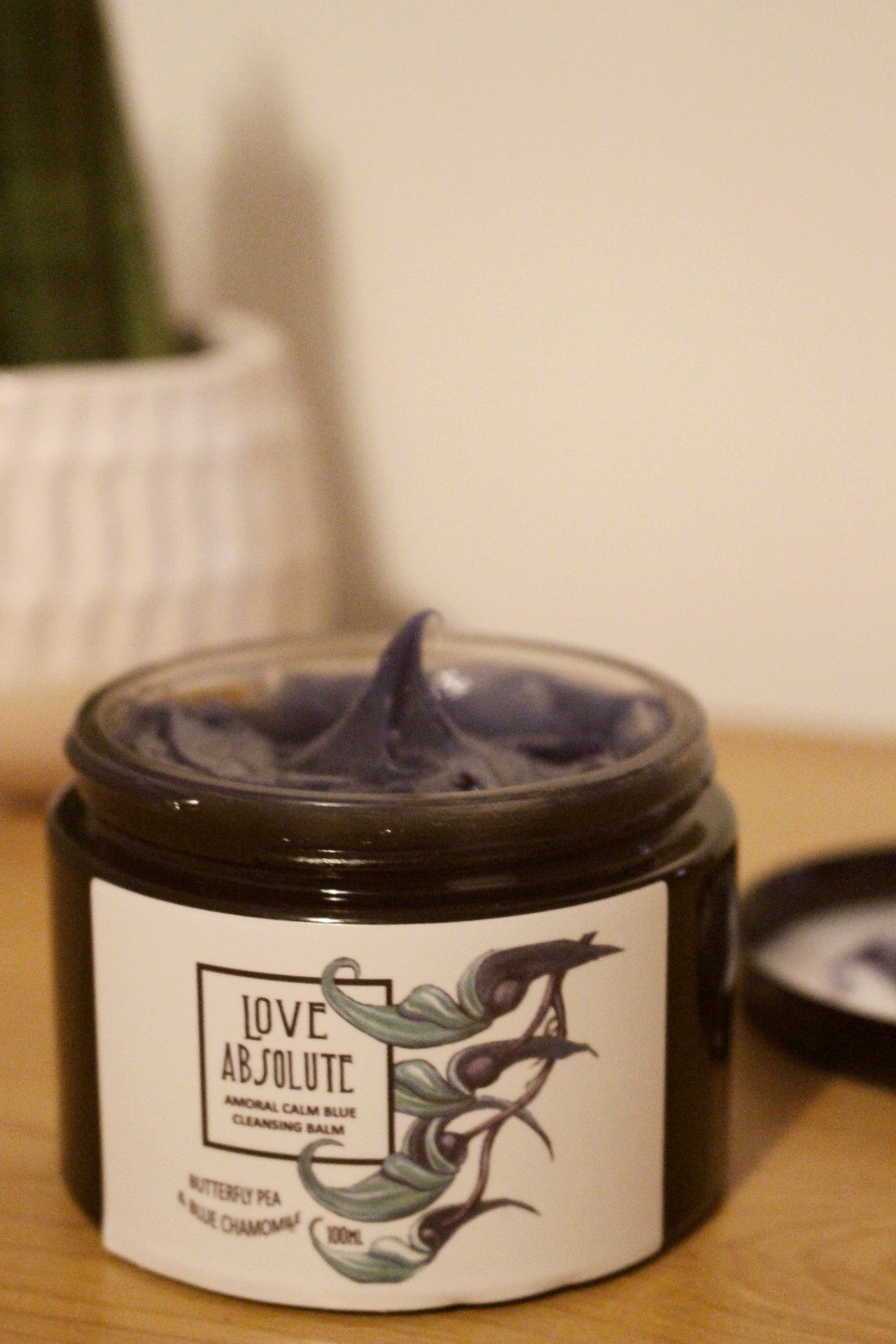 A dark coloured jar sat on a brow wooden surface. The jar has its lid removed and you can see a blue-purple product inside. The label on the jar reads "Love Absolute Amoral Calm Blue Cleansing Balm"