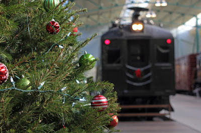 A Christmas tree with round ornaments and strings of lights is to the left. To the right, further in the background is one end of a railway passenger car