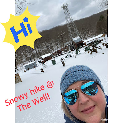 A woman with blond hair, a gray knit cap, and blue mirrored sunglasses takes a selfie. Behind her is snow-covered ground and an array of small buildings and oil field equipment, including a tall metal tower. A yellow starburst with "Hi" in blue letters and "Snowy hike @ the Well!" in red letters are added to the photo.