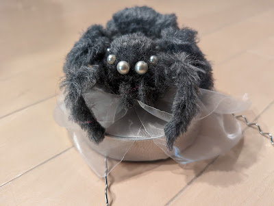 photo of tiny silver hat with a layer of sheer white fabric. A handmade fuzzy gray spider with pearl bead eyes sits on top of that. This whole thing is attached to a headband