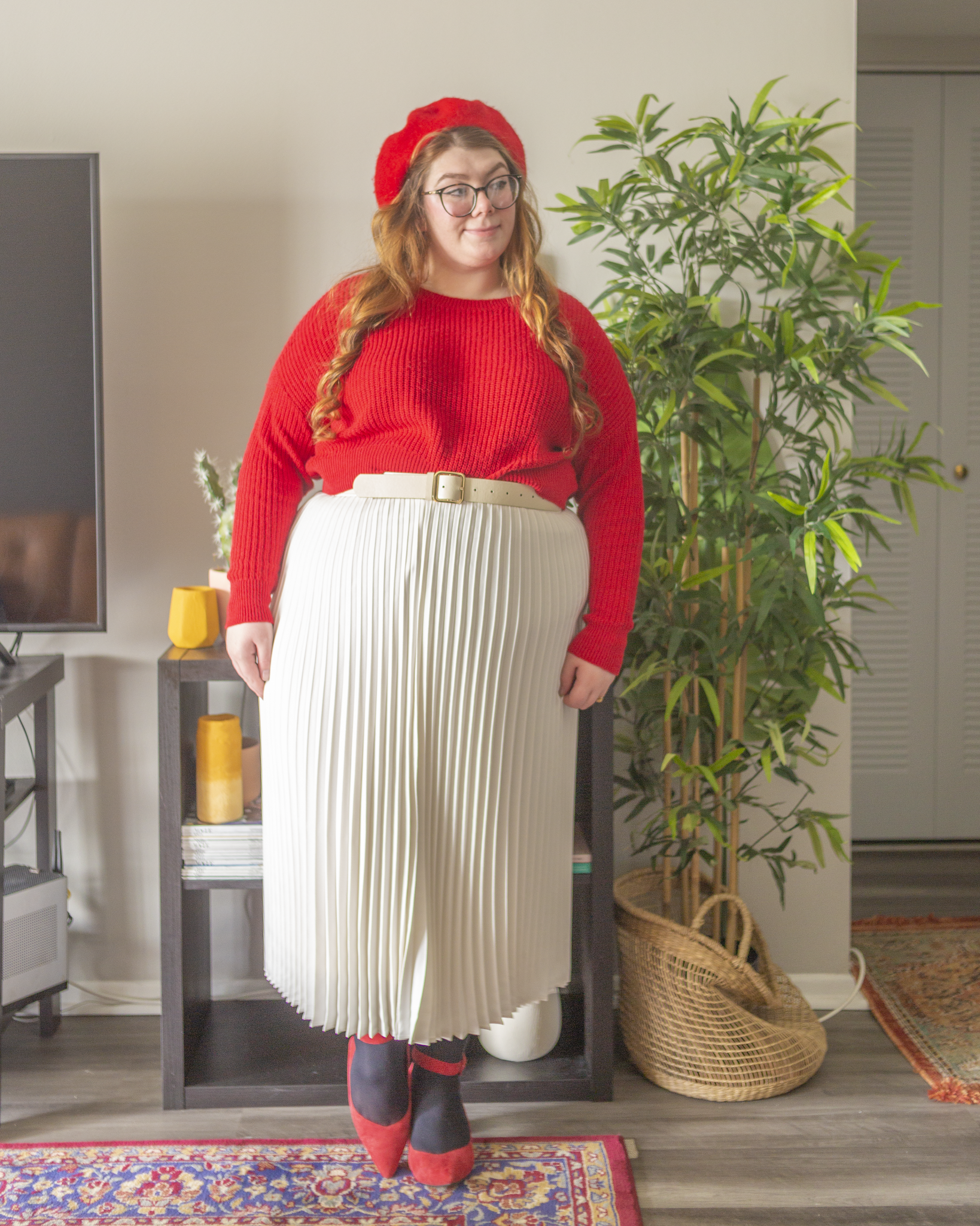 An outfit consisting of a red cropped sweater half tucked into a white pleated midi skirt and red ankle strap heels.