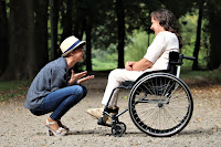 Caregiving on knees in front of wheelchair bound individual, smiling, having a conversation and making them laugh