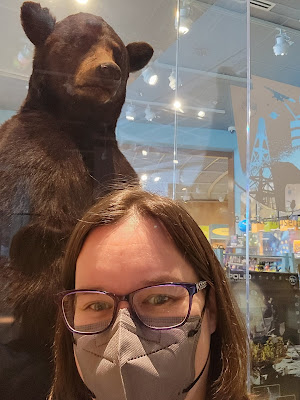 A woman with straight brown hair, glasses, and a gray mask takes a selfie. Behind her is a glass case with a taxidermied bear. Items in the museum store are also visible through a glass wall behind her.