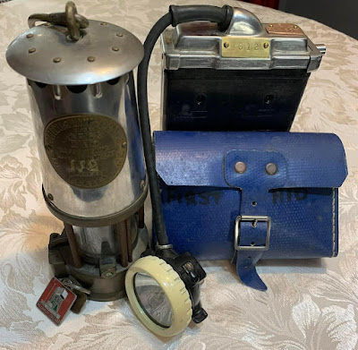 A tall cylindrical oil lamp, a black battery-powered lamp with the lamp on the end of a thick cable, and small blue leather satchel with 'first aid' written on it