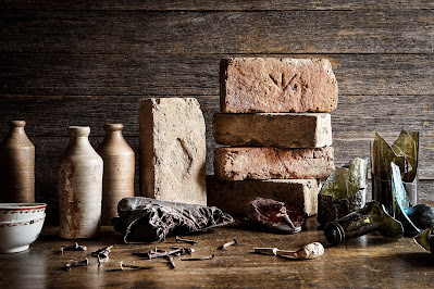 Historical artefacts on a table, consisting of bottles, broken glass bottles, nails, a shoe, a cup, and stacked bricks with designs etched into them