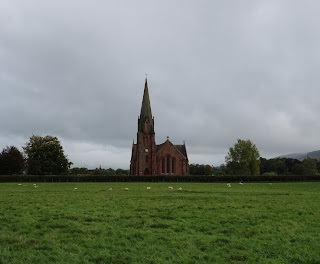 A photograph showing a view over a field to a church with a very tall spire in comparison to the building.  This is the Parish Church of Penpont, Keir and Tynron.  Photo by Kevin Nosferatu for the Skulferatu Project.