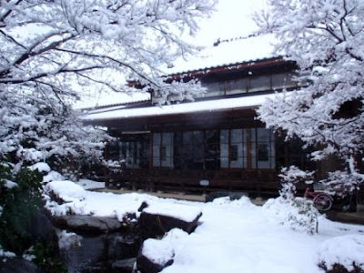 A snow-covered Japanese house seen from a garden with a pond.