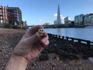 A picture of a small ceramic skull (Skulferatu #56) being held up.  In the background can be seen the foreshore of the River Thames and in the distance the Shard building.  Photograph by Kevin Nosferatu for the Skulferatu Project