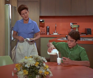 Greg pours milk in the kitchen while Alice questions him about what he just said.