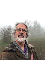 head shot of John in a brown jacket standing in front of fog-obscured oaks and pines.