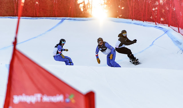 Três snowboarders contornam curva de circuito de snowboard cross. A pista na neve é demarcada por linhas azuis e bandeiras vermelhas