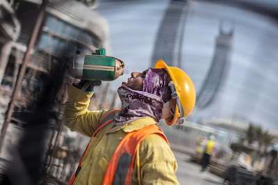 Color photograph of a worker in construction gear drinking from a water bottle on a World Cup construction site in Lusail City, Qatar to stay hydrated during extreme heat conditions.