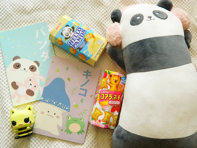 A photo showing a collection of cute christmas gifts: a cat-bee funko pop figure, two boxes of japanese chocolate snacks, two notebooks with panda and frog covers, a panda plush wearing earmuffs