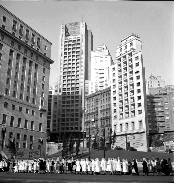 A fotografia mostra um lugar com vários prédios em volta. Também há a presença de muitas pessoas, algumas delas parecem ser freiras, caminhando na rua.