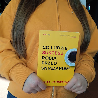 Co ludzie sukcesu robią przed śniadaniem, książkę Laury Vanderkam prezentuje czytelniczka z długimi ciemnymi włosami, ubrana w słoneczną bluzę.