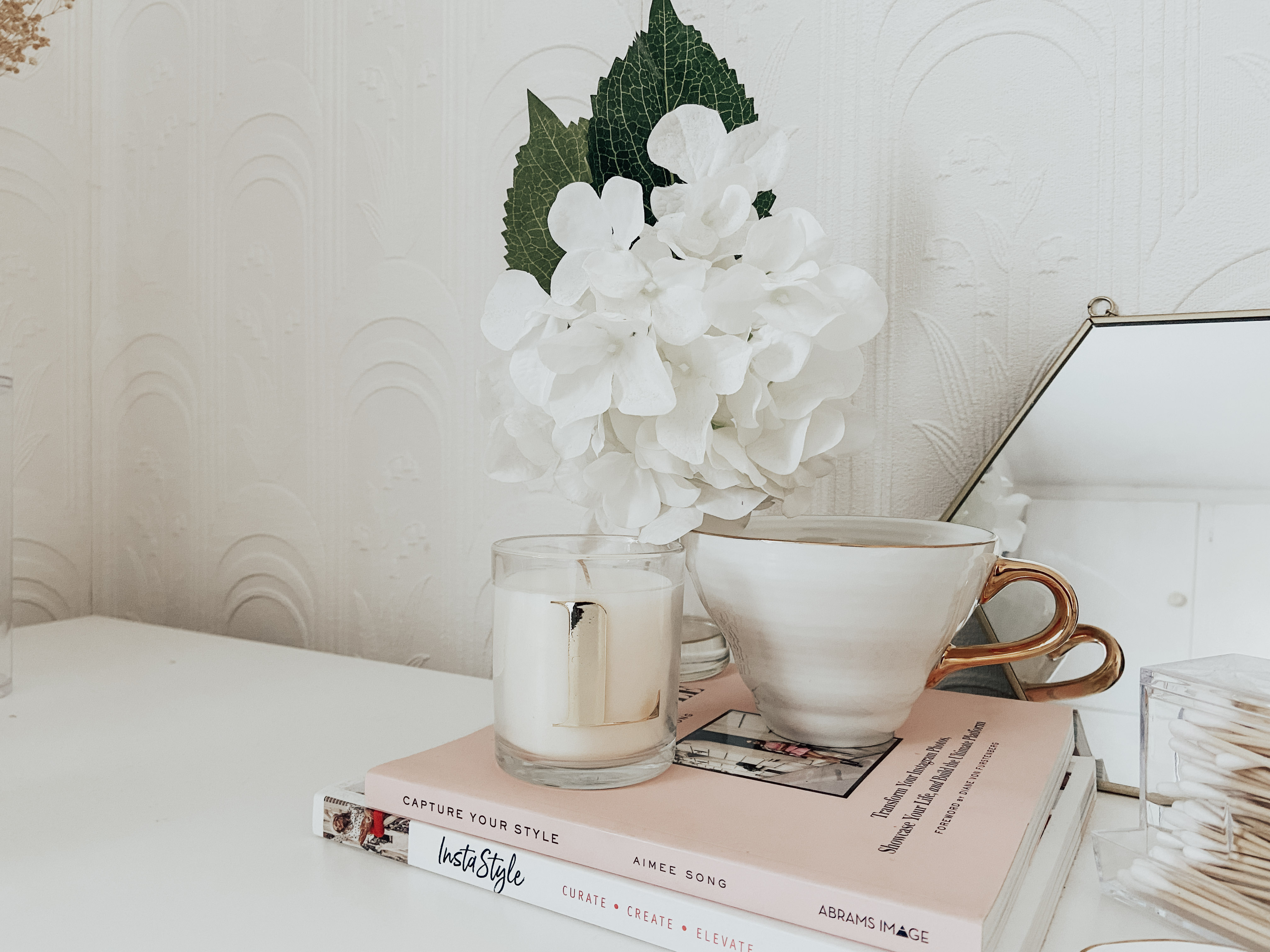 A white mug and white artificial plant on top of pink books.