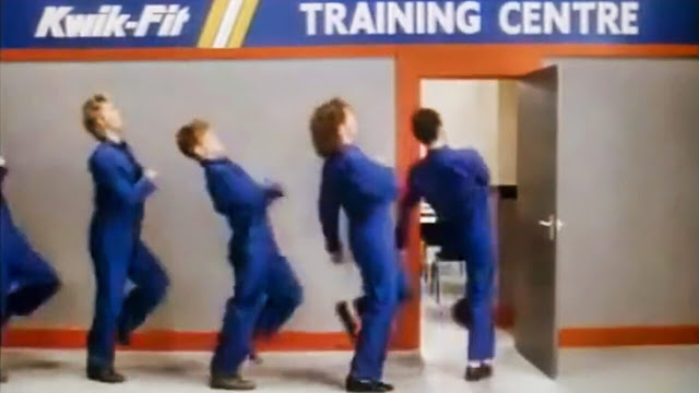 A group of men in blue overalls dancing in line at a Kwik Fit Training Centre