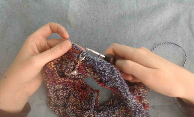 Someone crocheting a crochet cowl in a shell star stitch, with variegated yarn.  There is a stitch marker with a shell charm clipped into the work.