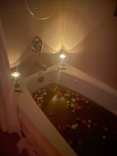 Bathtub filled with brown water made from dried flowers and herbs with orange, pink and white roses floating. Candles lit on corners of tub