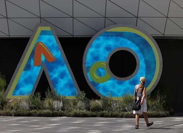 Mulher loira de vestido branco passa na frente do símbolo do Australian Open, que é formado pelas letras A e O preenchidas em azul, com detalhes em verde marca texto claro