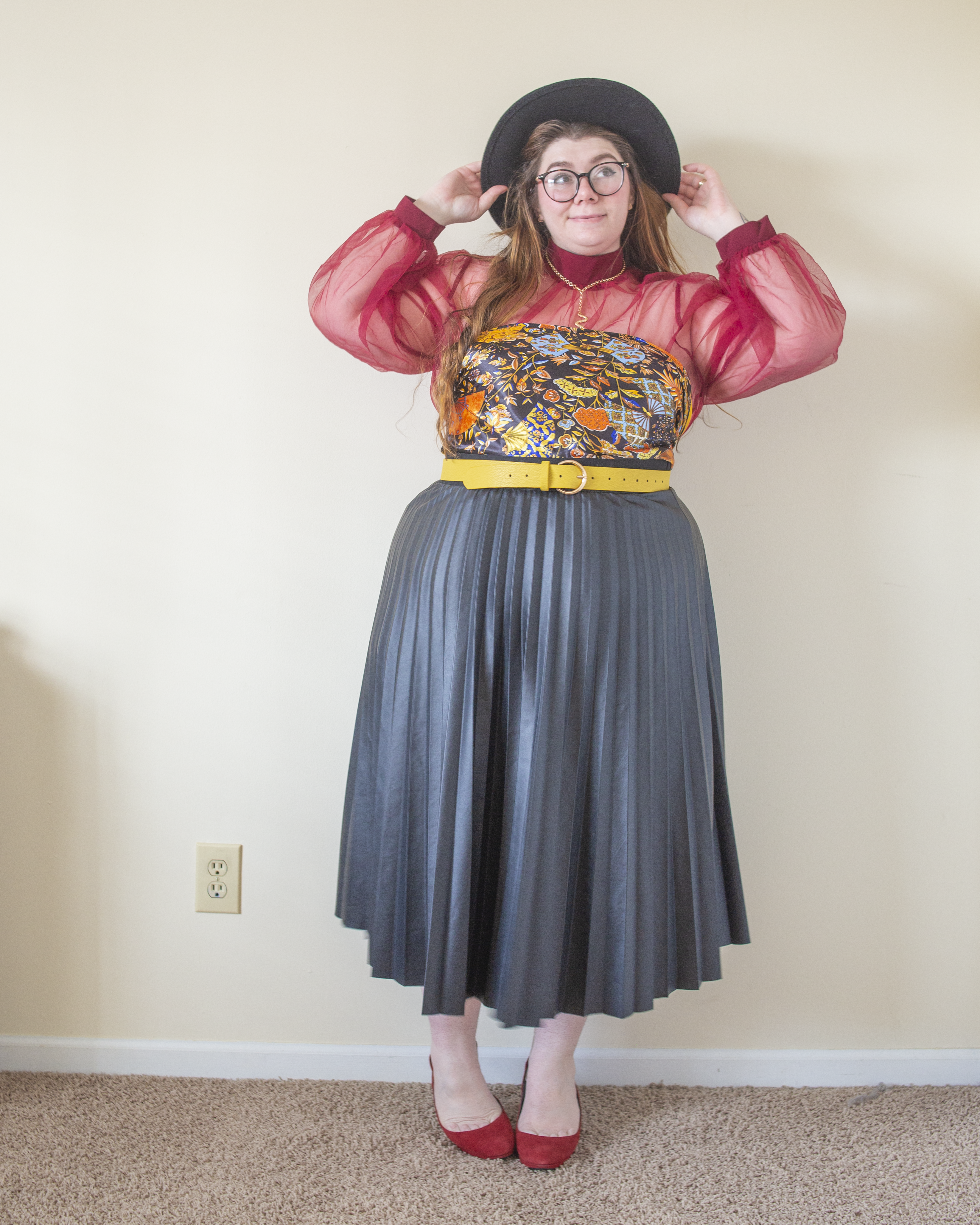 An outfit consisting of a sheer red blouse with lantern sleeves with a black satin scarf with a navy blue, light blue, gold and orange floral motif tied as a top layered over, a black leather pleated midi skirt and red round toe cling back heels.