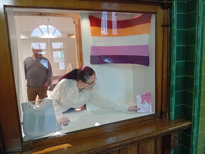 Fern leaning inside the glass display case in the Pontefract Museum foyer, placing a copy of the Grayson's Art Club exhibition catalogue in the window. Their crocheted Lesbian Pride flag is hung up to the right of the case.