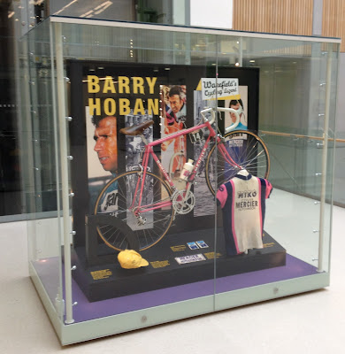 Square glass display case featuring photos and objects relating to Barry Hoban's career, including a pink road bike and pink, white and blue jersey