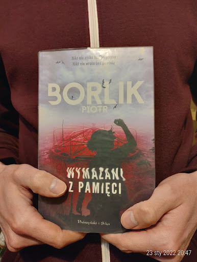 Mężczyzna w bordowej bluzie z białym zamkiem prezentuje książkę Piotra Borlika pod tytułem Wymazani z pamięci.