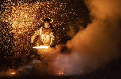 color photograph of an ironmonger in protective gear spinning molten iron while fellow artists throw liquid iron onto a plywood wall behind him that causes a cascade of yellow sparks