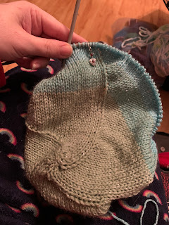 The blanket is now on a circular needle and this phot shows the progression of the yarn's colors from mossy green to light blue, and the transition section between them. There's a silver dangling heart shaped stitch marker on the circular needle, visible near the top of the photo