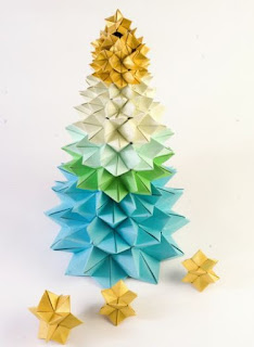 Foto em fundo branco de uma árvore de Natal composta por dezenas de poliedros estrelados encaixados e bem arranjados feitos por dobraduras de papel(Origami). No topo, em tom dourado, em seguida, branco, verde e finaliza em azul.  Próximo da árvore, no chão branco, três peças soltas em forma de estrelas douradas.  Desejamos um Feliz Natal a todos!