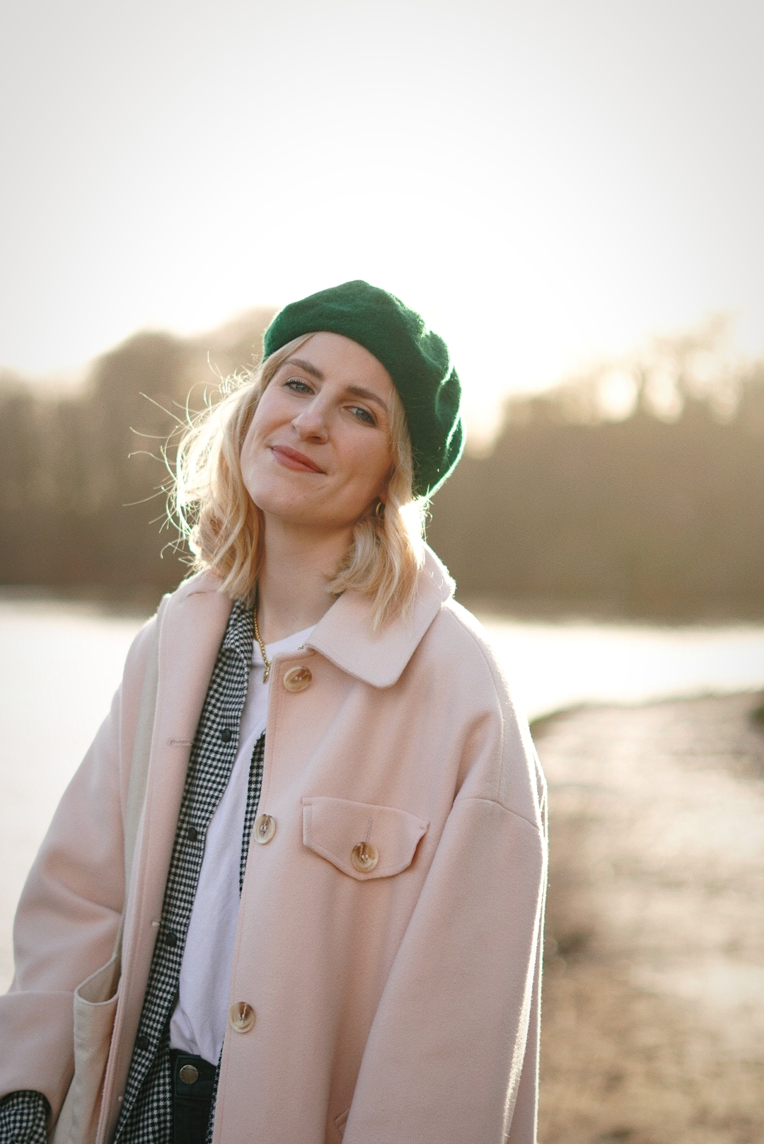 Amy is smiling at the camera, wearing a green beret and a light pink coat. She is stood in front of a lake, the sun casting a warm glow over the photo.