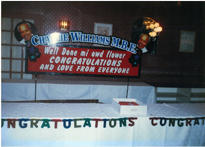 Decorated table and hall for Charlie Williams' MBE party. There is a banner on the wall reading 'Charlie Williams M.B.E. Well Done mi owd flower. Congratulations and Love from Everyone!'. The banner is flanked by two photos of Charlie