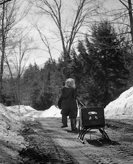 black and white photograph of young child pulling a sled behind them on a dirt road with snowy embankments