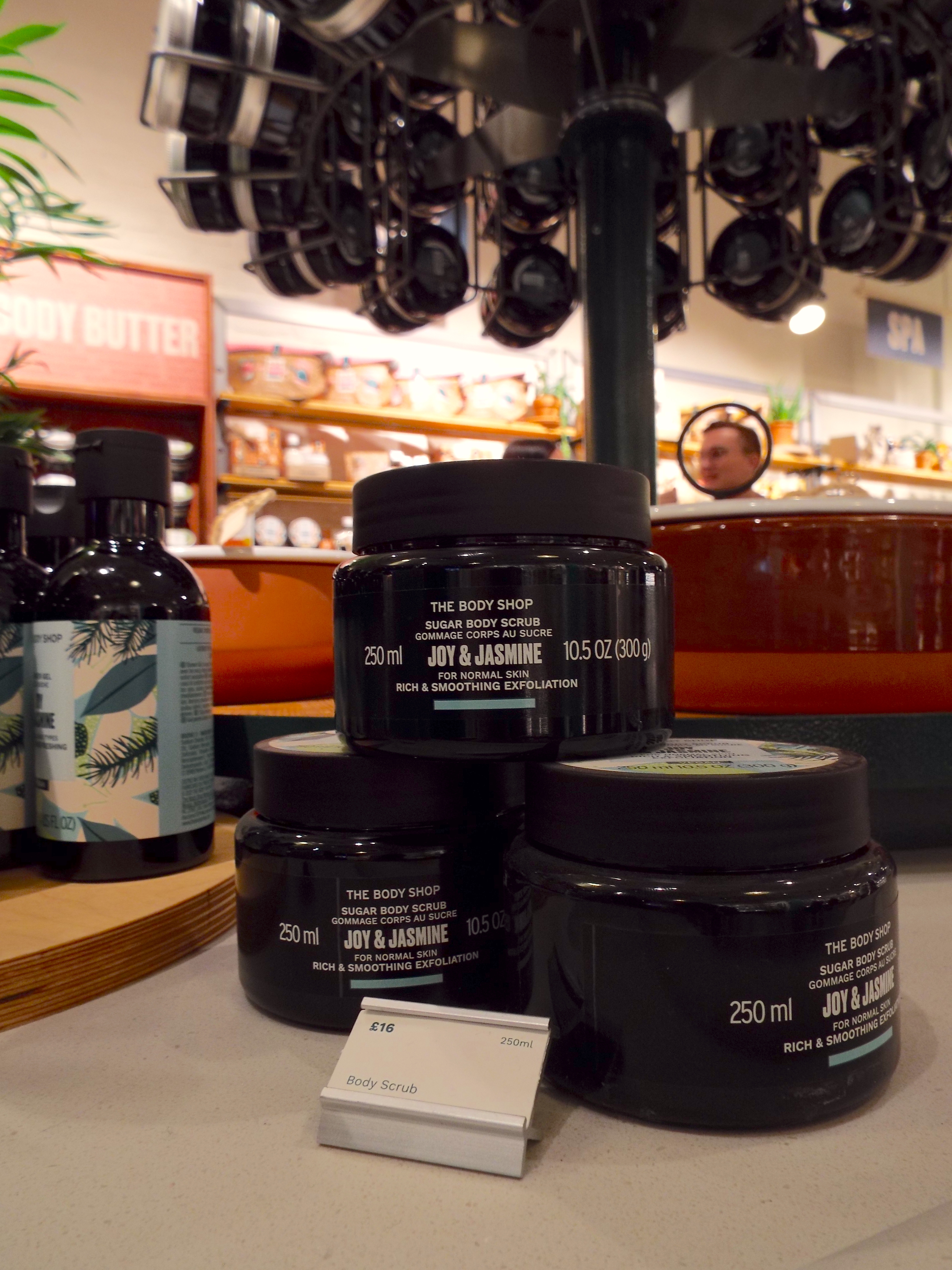 A close up shot of a stack of three Joy and Jasmine Body Scrubs, with Ryan stood in the background but his little head is haloed with a piece of the display, like a little angel.