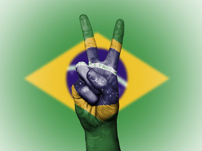 Bandeira do Brasil, projetada sobre mão fazendo sinal de paz e amor. #PraCegoVer