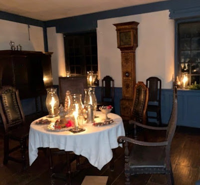 A room is set with heavy 17th-century chairs and other furniture. A round table in the middle of the room has a white cloth and is covered with dishes of food and candles with glass hurricane lanterns