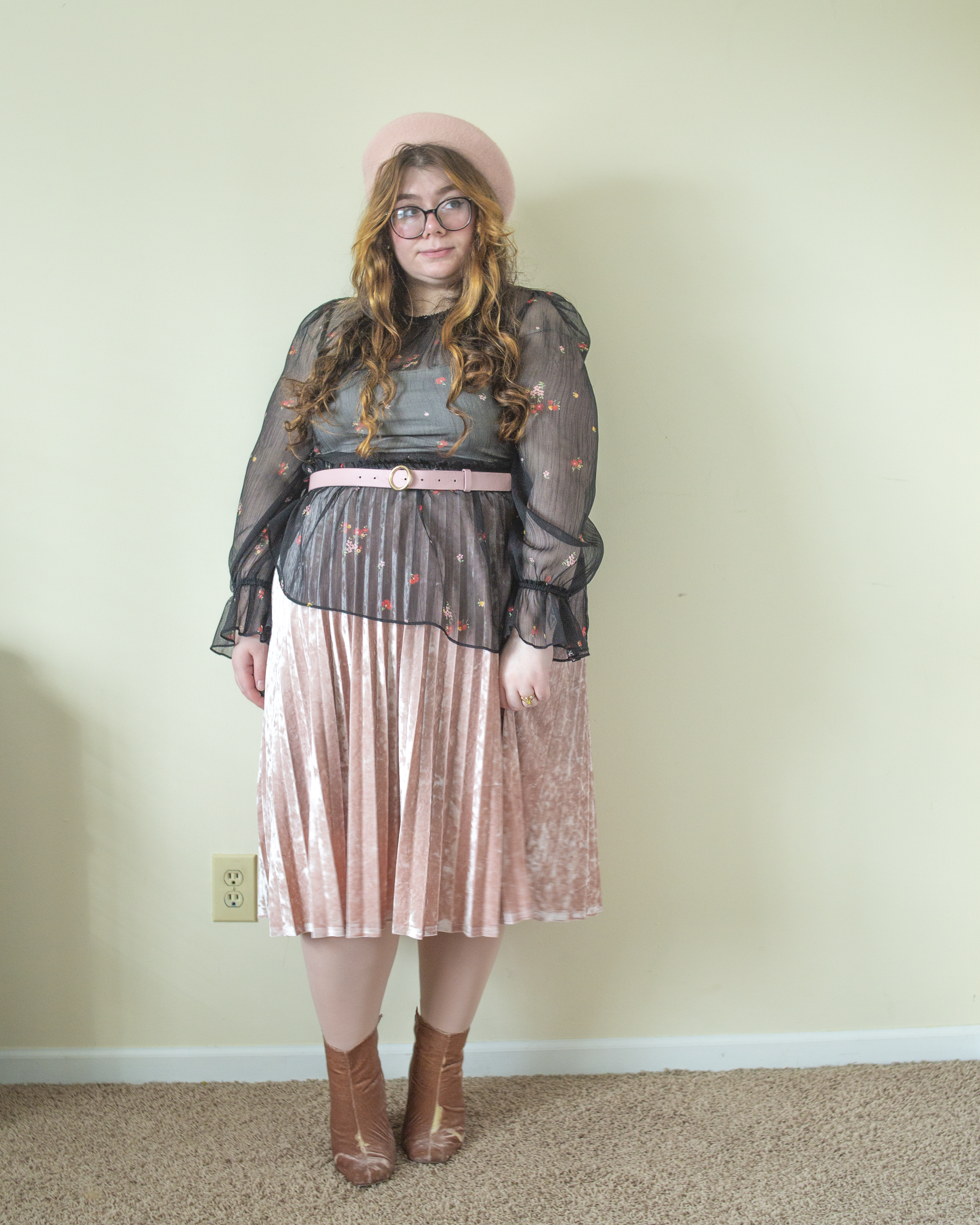 An outfit consisting of a pastel pink beret, a black sheer long bishop sleeve dress with micro floral print with asymmetrical tiers over a pastel pink velvet pleated midi skirt and pastel pink velvet midi boots.